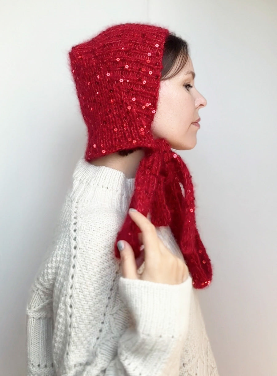 Woman wearing a handmade red knitted bonnet with sequins, inspired by Little Red Riding Hood. Cozy winter accessory perfect for Halloween costume or cottagecore fashion.
