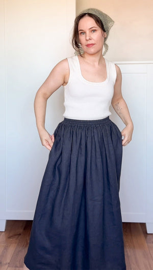 Woman wearing a white tank top and navy blue skirt standing against a white wall.