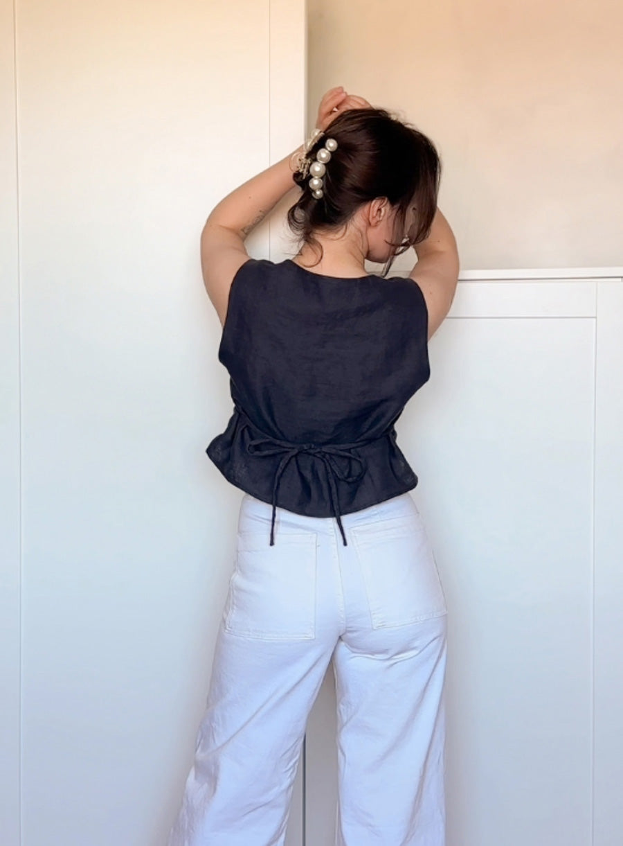 Back view of a woman wearing a sleeveless black linen top with a tie closure at the waist, paired with high-waisted white pants