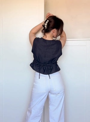 Back view of a woman wearing a sleeveless black linen top with a tie closure at the waist, paired with high-waisted white pants