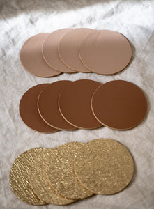 A set of round coasters in three different colors—light beige, dark brown, and textured gold—are neatly arranged on a soft, neutral-colored fabric surface. Each group of coasters is stacked slightly overlapping, showcasing their smooth and textured finishes. The gold coasters have a glittery, rough texture, while the beige and brown ones have a matte, leather-like appearance.
