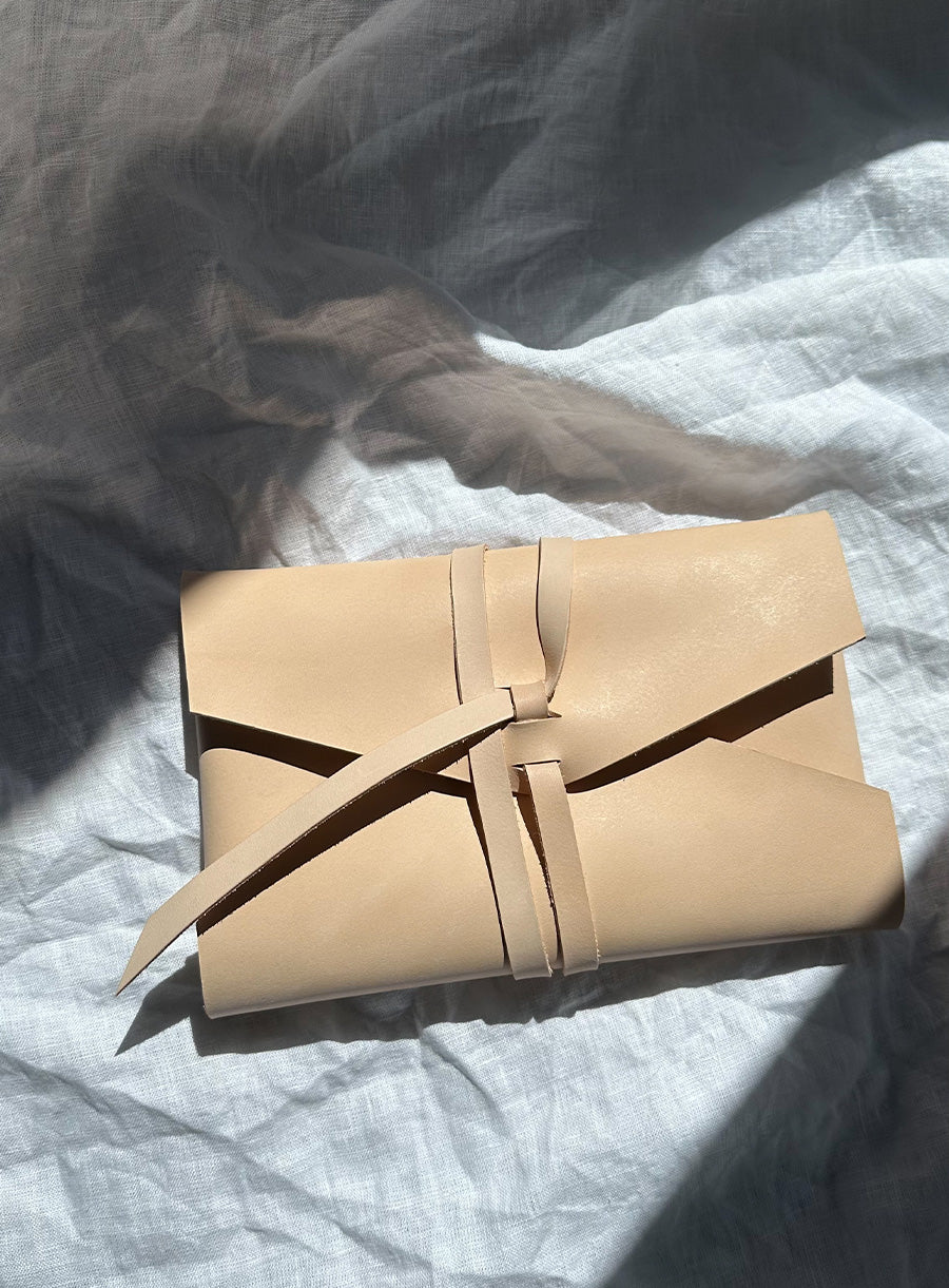 A beige leather pouch with a wraparound strap closure is placed on a softly wrinkled white linen fabric. Sunlight casts dappled shadows across the fabric, creating a warm and natural aesthetic.