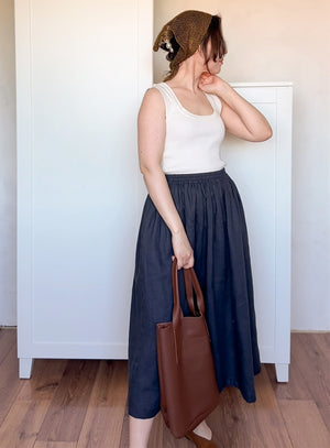 Woman wearing a white top and navy skirt, holding a brown leather bag , and linen bandana