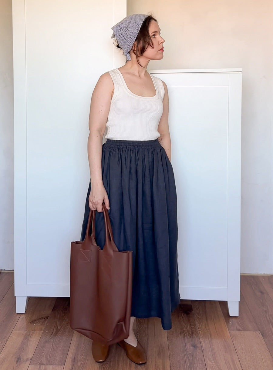 Woman wearing a white tank top and navy skirt, holding a brown bag and pastel blue head scarf
