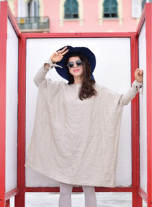 A stylish woman poses in an oversized beige linen dress with flowy wide sleeves. She pairs it with white leggings, a navy blue floppy hat, and round sunglasses. She makes a playful peace sign with one hand, standing against a red-framed structure with a pastel pink and green building in the background