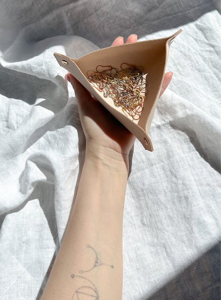 Hand holding a beige leather catchall tray filled with gold and silver stitch markers, against a soft white linen background