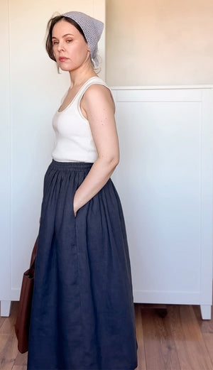Woman wearing a white tank top and navy blue skirt with a baby blue beanie indoors.