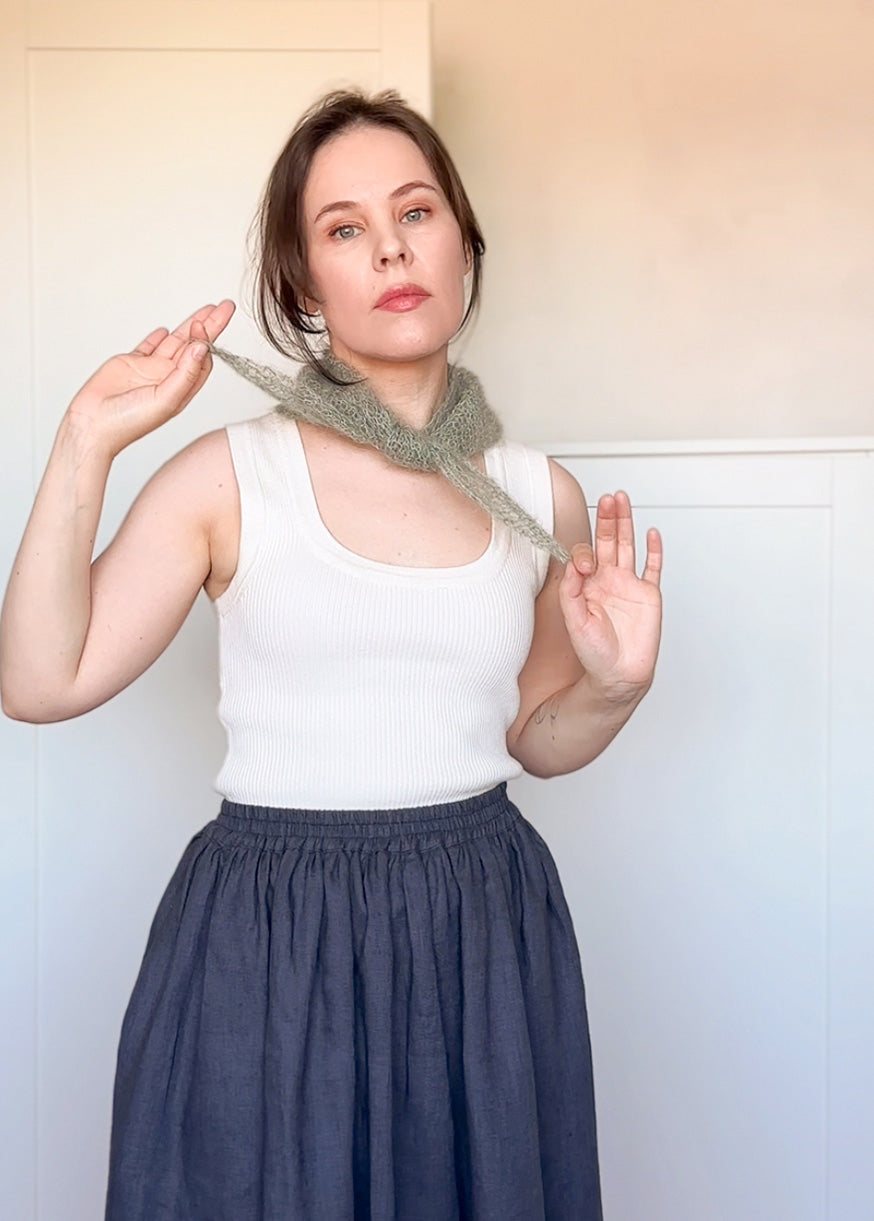 Woman wearing a white tank top and dark blue skirt with a green knitted scarf.