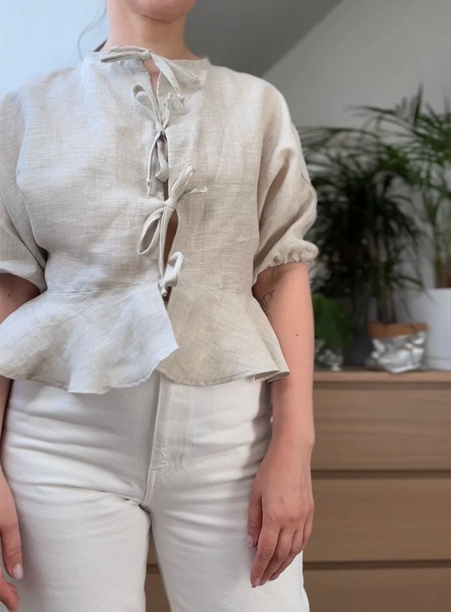 Women's linen blouse in a natural beige color with a delicate peplum hem. Features short puffed sleeves with elastic cuffs and a unique front closure with three adjustable bow ties. Lightweight and breathable, perfect for an effortless, chic look. Styled with high-waisted white pants. Ideal for casual or semi-formal wear