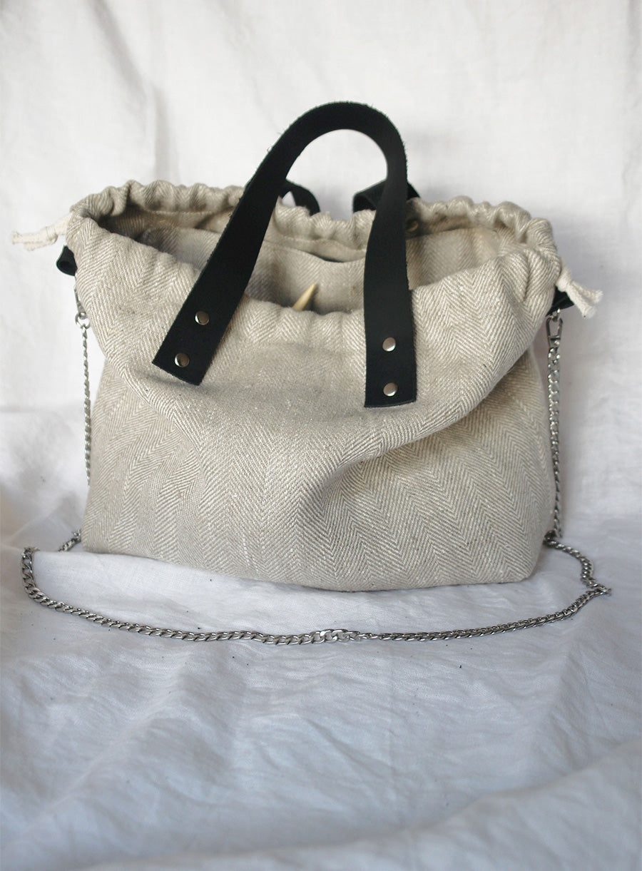 Linen knitting project bag made from high-quality heavy linen fabric, featuring a stylish herringbone pattern. Designed with sturdy black leather handles, a secure drawstring closure, and a silver chain strap for versatile carrying options. Perfect for storing and transporting knitting or crochet projects with elegance and durability