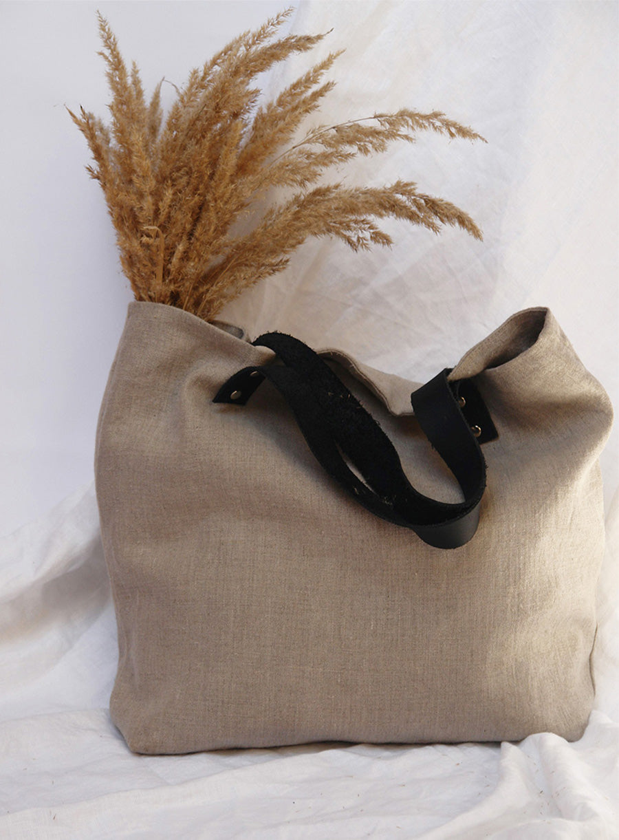 Minimalist linen tote bag with black leather handles and dried pampas grass, set against a neutral fabric backdrop.