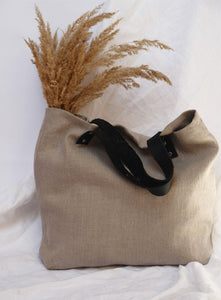 Minimalist linen tote bag with black leather handles and dried pampas grass, set against a neutral fabric backdrop.