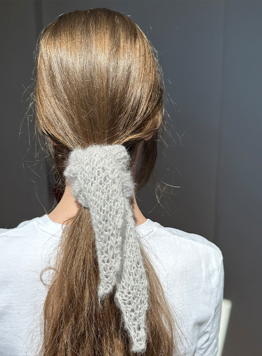 Person with long brown hair tied into a ponytail with a white knitted scarf, wearing a white sweater against a dark background.
