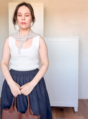 Woman wearing a white sleeveless top and blue skirt, holding a brown bag indoors.