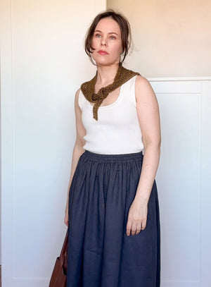 Woman wearing a white sleeveless top and navy skirt with a green patterned scarf, standing against a plain background.