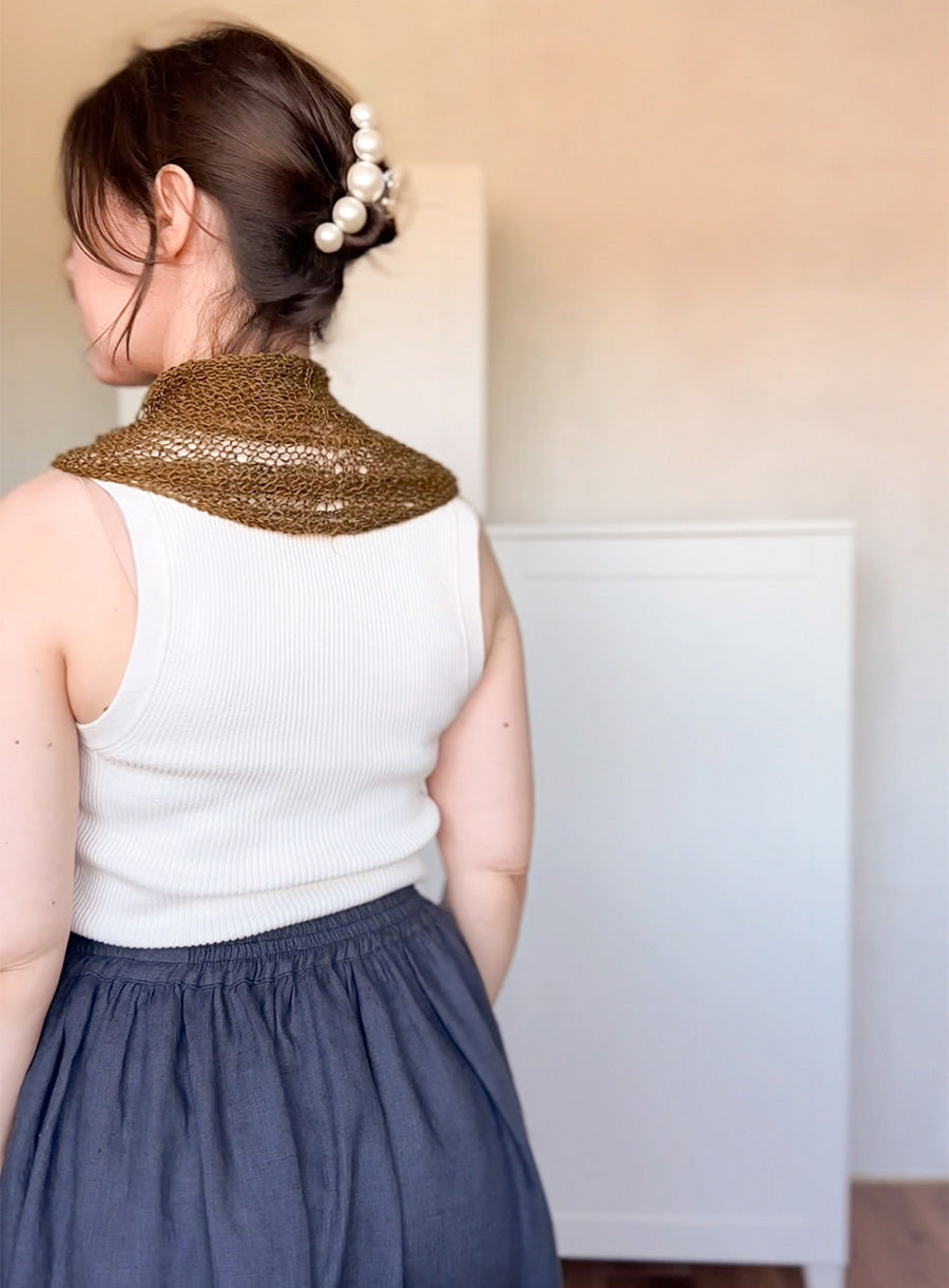 Woman wearing a white sleeveless top and blue skirt with a green linen yarn knitted shawl over her shoulders, standing against a plain background.