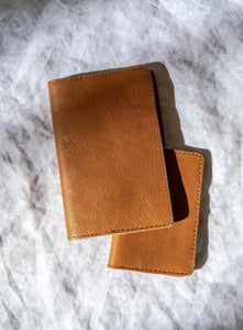 VEGETABLE TANNED leather passport holders resting on a textured white fabric surface with natural sunlight casting soft shadows. The leather has a smooth, slightly worn texture with fine stitching along the edges