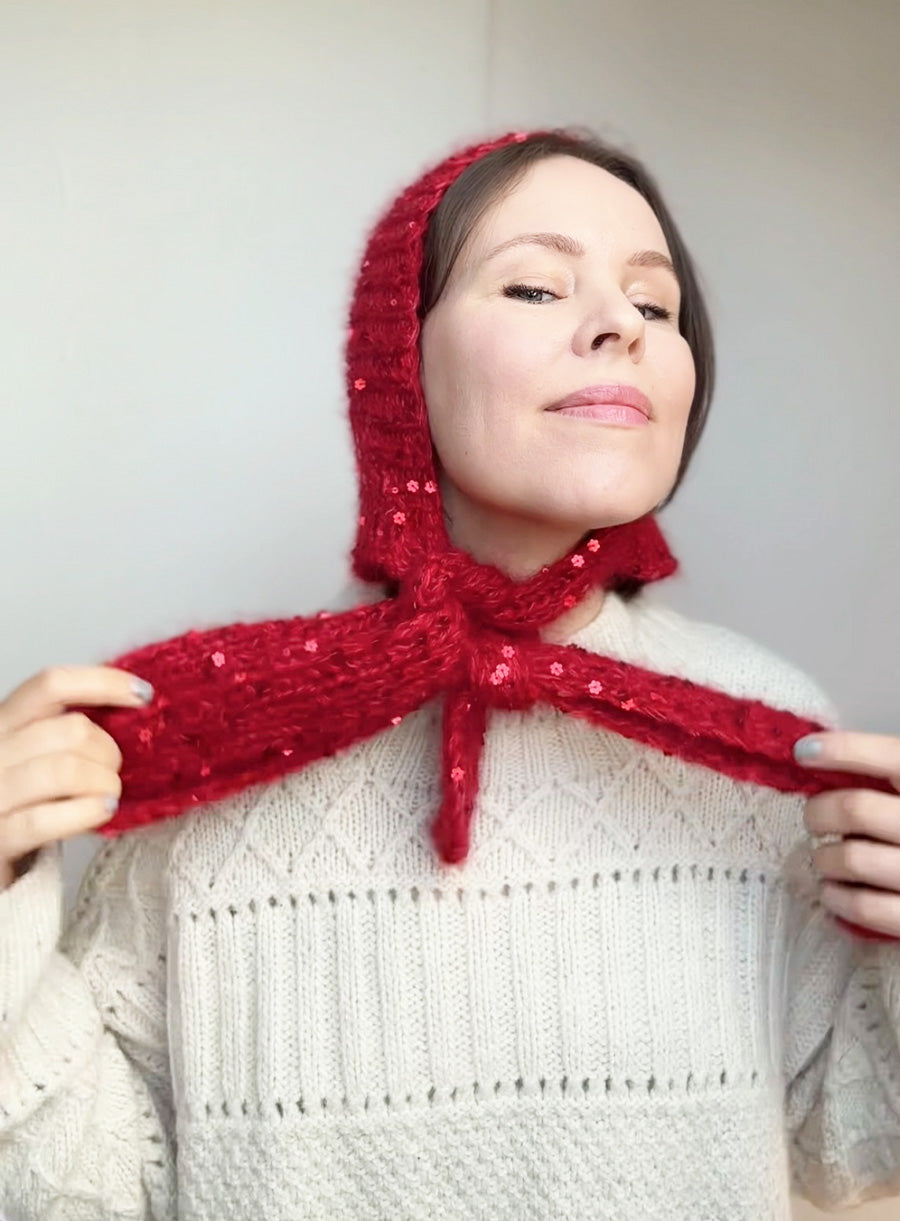 Stylish red knitted bonnet with a large tie under the chin, worn by a woman in a textured white sweater. Ideal for cottagecore or vintage-inspired styling.