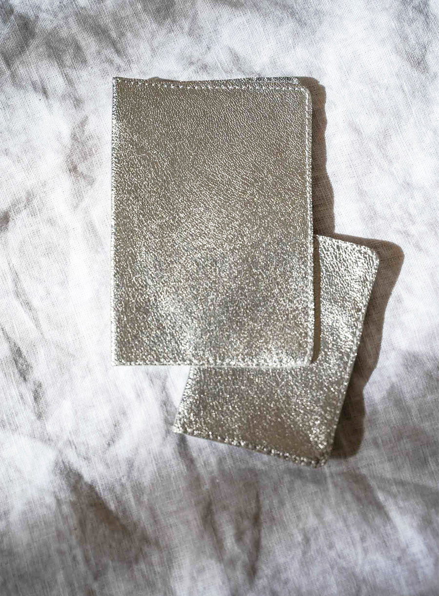 Two tan-colored leather passport holders resting on a textured white fabric surface with natural sunlight casting soft shadows. The leather has a smooth, slightly worn texture with fine stitching along the edges.
