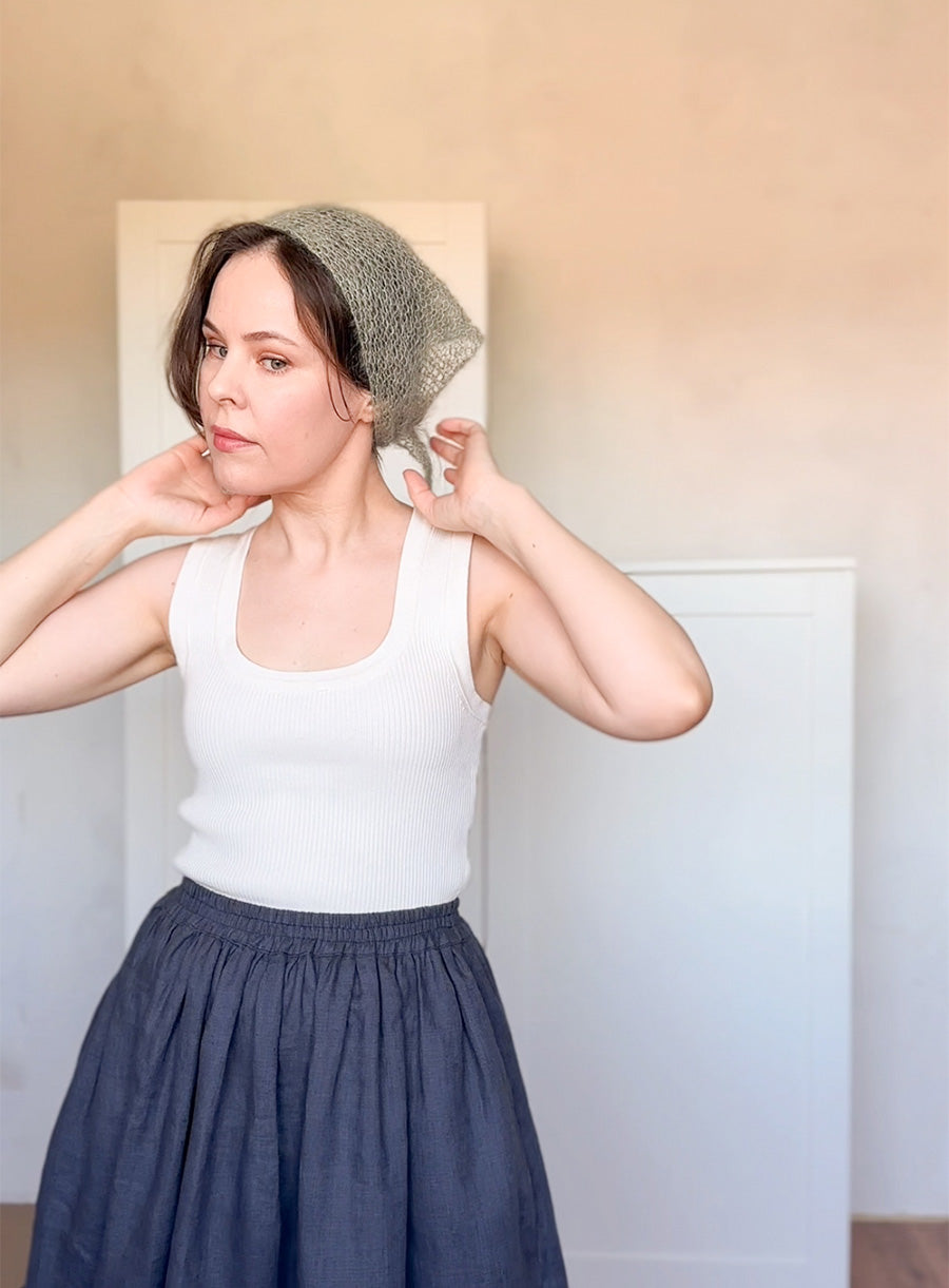 Woman wearing a white tank top and blue skirt with a textured headscarf indoors.