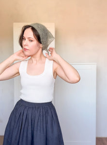 Woman wearing a white tank top and blue skirt with a textured headscarf indoors.