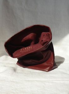 Burgundy red leather clutch bag with a folded design, placed on a white fabric background in natural sunlight