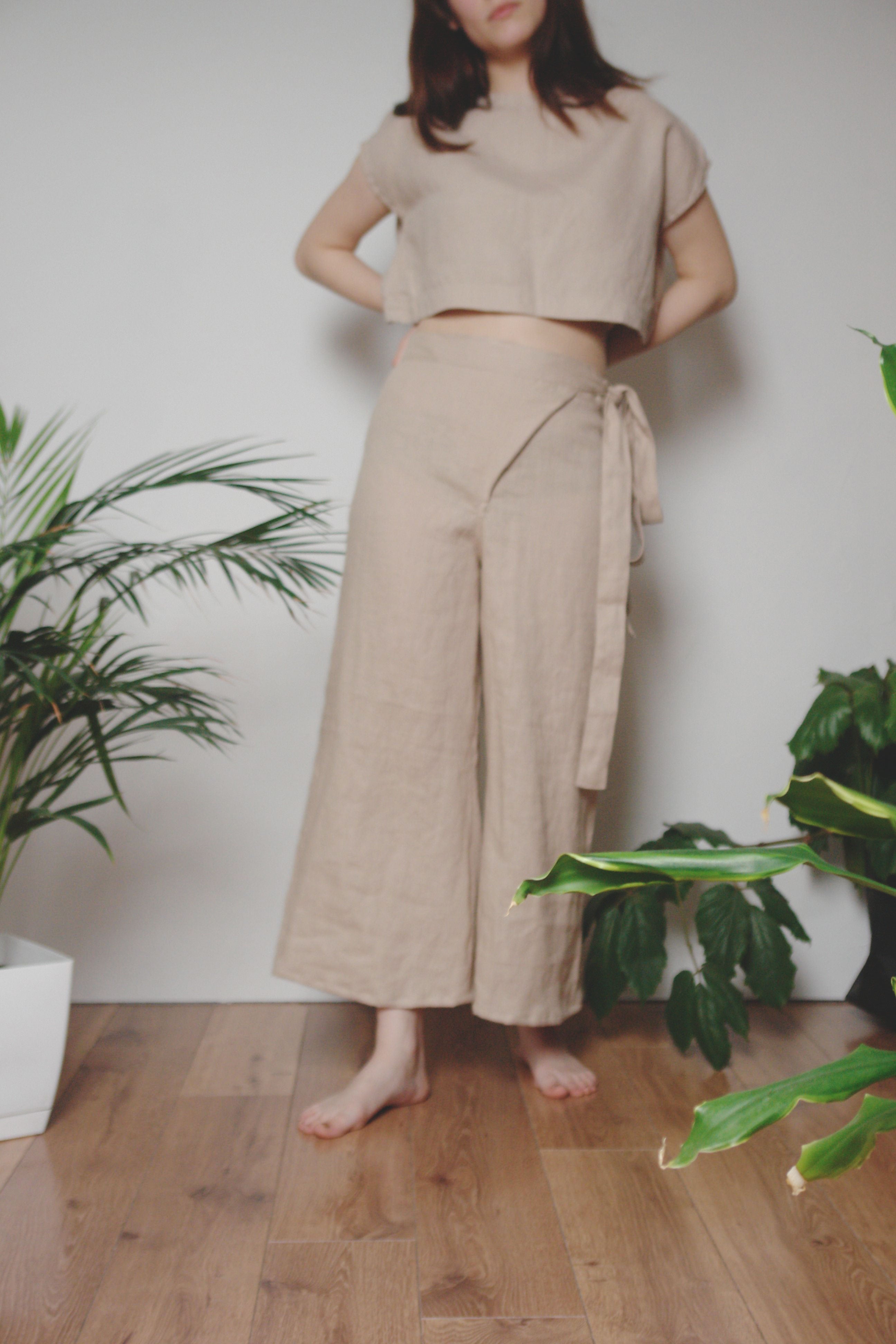 Stylish beige linen two-piece outfit with a cropped top and wrap pants, exuding a relaxed, minimalist aesthetic. The lightweight fabric and neutral tones create a breathable, effortless look. The model is barefoot, standing in a cozy indoor setting with wooden flooring and lush green plants, enhancing the natural and organic vibe